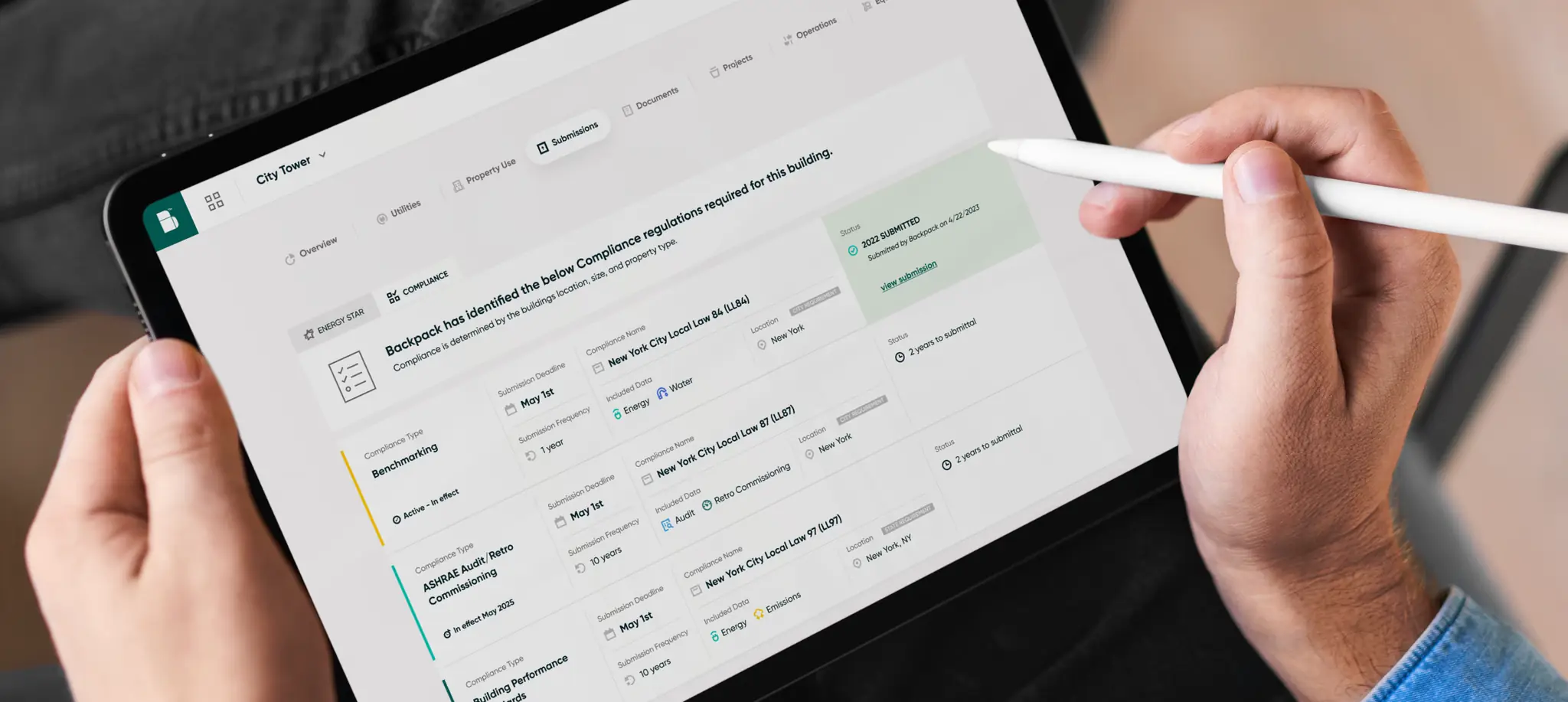 Hands holding a tablet with a stylus, the screen showing the compliance regulations Backpack has identified for a building
